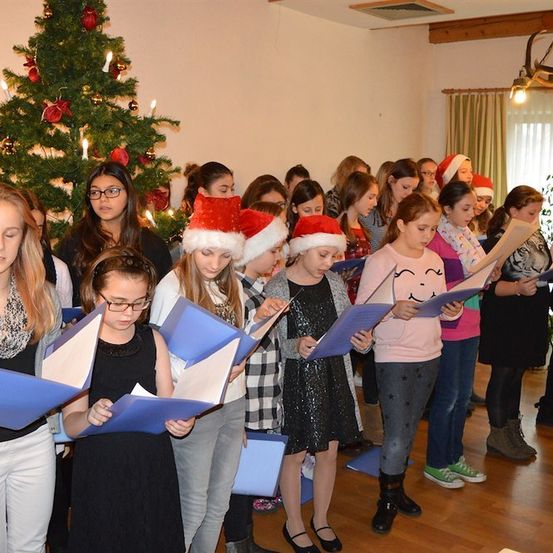 Eine Gruppe von Kindern in festlicher Kleidung singt Weihnachtslieder und hält Notenblätter in einer für Weihnachten geschmückten Stube.