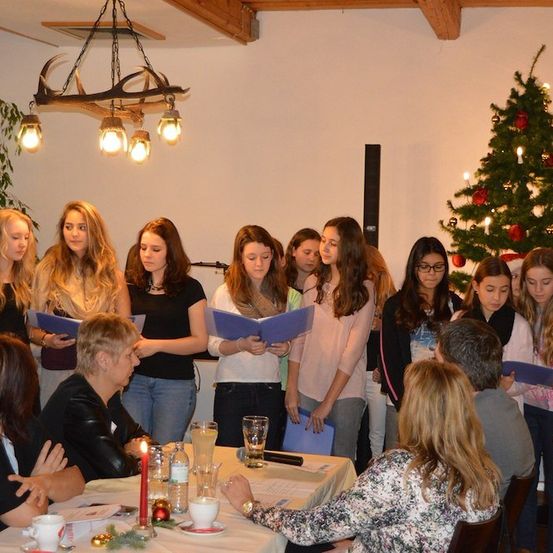Eine Gruppe junger Frauen und einige Erwachsene stehen in einem Raum, halten blaue Hefte und singen. Ein Tisch mit einer Kerze, Tassen und einem Teller steht vor ihnen. Ein Weihnachtsbaum befindet sich auf der rechten Seite.