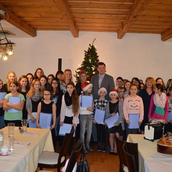 Eine Gruppe junger Mädchen und Frauen steht in einem Raum mit einem Weihnachtsbaum und hält Urkunden in den Händen, während sie für ein Foto lächeln. Ein Mann in einem Anzug steht vor ihnen.