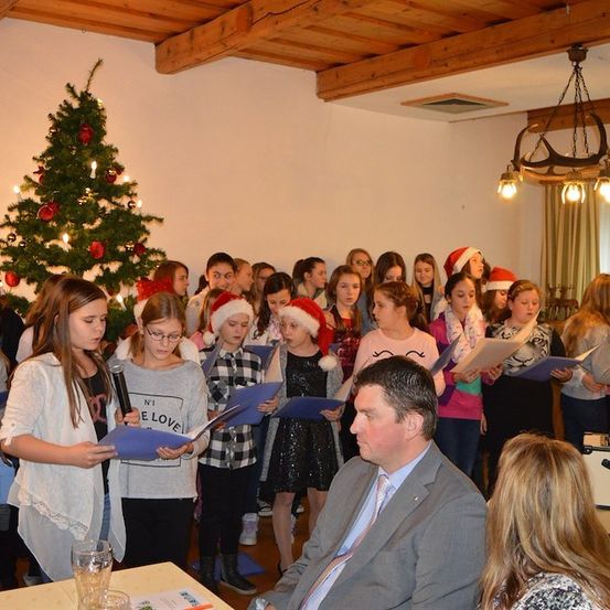 Eine Gruppe junger Mädchen und Jungen singt Weihnachtslieder mit einem Mann in Anzug vor ihnen. Sie halten Papiere und tragen Weihnachtsmützen.