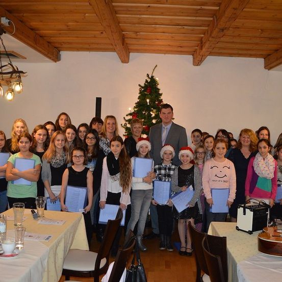 Eine Gruppe von Menschen, einschließlich Kindern, versammelt sich in einem Raum mit einem Weihnachtsbaum und Dekorationen. Sie halten Urkunden und posieren für ein Foto.