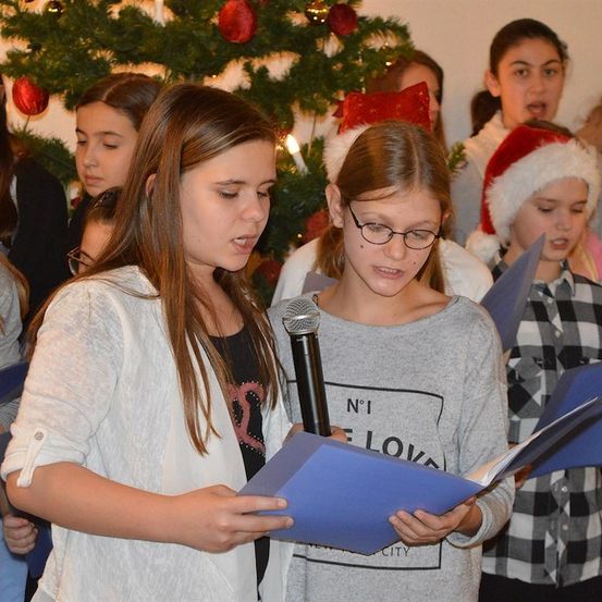 Eine Gruppe junger Mädchen in festlicher Kleidung, die blaue Papiere halten und vor einem geschmückten Weihnachtsbaum singen.