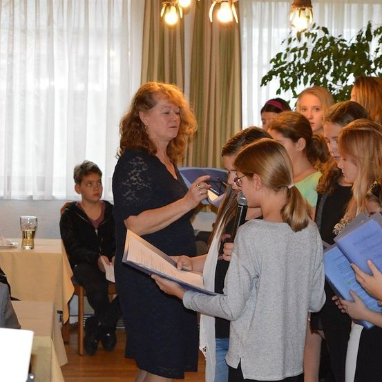 Eine Frau mit Mikrofon und Buch steht vor einer Gruppe junger Mädchen, die ebenfalls Bücher halten und singen. Ein Junge sitzt im Hintergrund an einem Tisch.