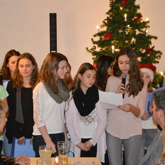 Eine Gruppe junger Mädchen steht vor einem Weihnachtsbaum, eine hält ein Mikrofon und liest von einem Stück Papier. Sie sind warm gekleidet, und eine Frau sitzt an einem Tisch in der Nähe.