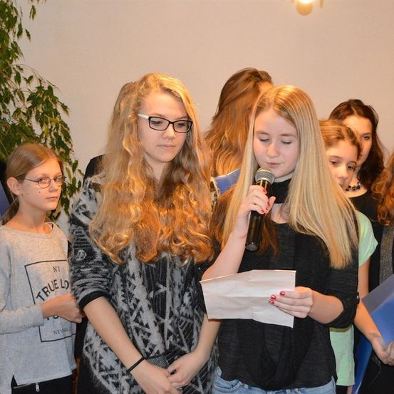 Eine Gruppe junger Mädchen mit blonden Haaren steht in einem Raum, eine von ihnen hält ein Mikrofon und ein Stück Papier. Sie sind wahrscheinlich bei einer Schulveranstaltung.