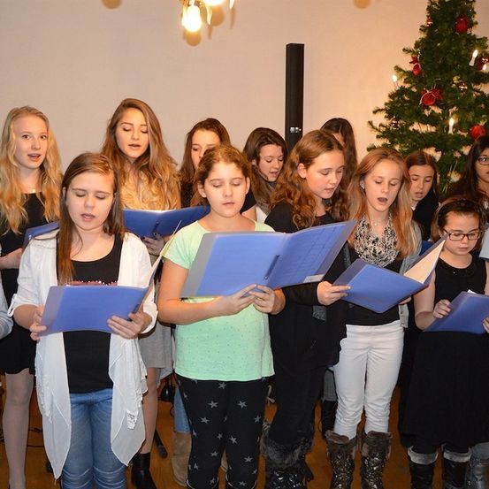 Eine Gruppe junger Mädchen singt in einem Chor, hält blaue Notenblätter in der Hand und steht vor einem Weihnachtsbaum.