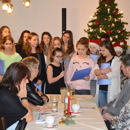 Eine Gruppe junger Mädchen und Frauen steht um einen Tisch, einige halten Papiere, andere ein Mikrofon. Sie sind für den Winter gekleidet, und ein Weihnachtsbaum steht hinter ihnen.
