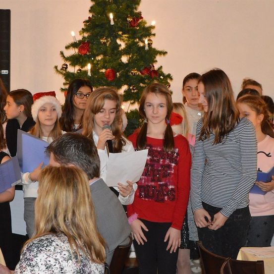 Eine Gruppe junger Mädchen steht vor einem geschmückten Weihnachtsbaum, hält Papiere in den Händen und singt, während ein Mann in einem Anzug in der Nähe steht.