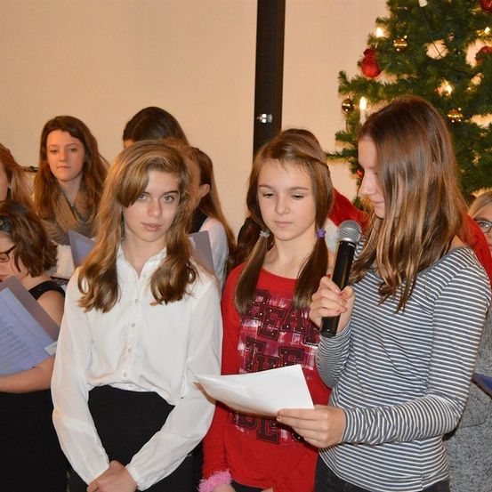 Eine Gruppe junger Mädchen steht in einem Raum, einige halten Papiere und andere halten ein Mikrofon, möglicherweise singen sie während einer Weihnachtsveranstaltung.