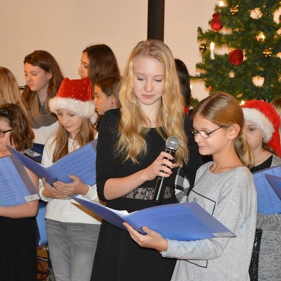 Eine Gruppe junger Mädchen, gekleidet in festlicher Kleidung, singen zusammen und halten blaue Liederbücher und ein Mikrofon. Das Mädchen in der Mitte trägt einen Weihnachtshut.