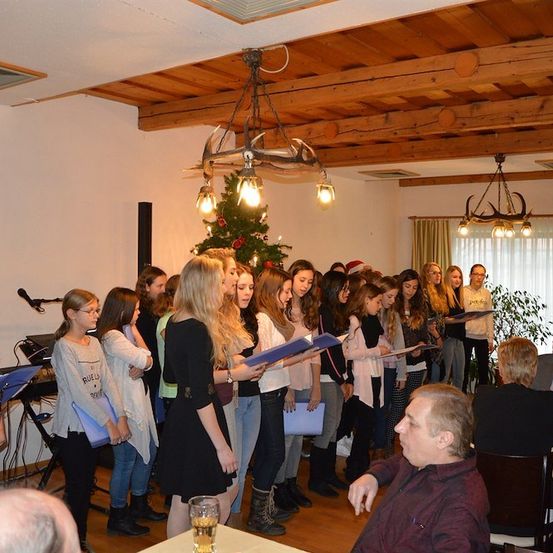 Eine Gruppe junger Frauen und Mädchen singt in einem Raum mit einem Weihnachtsbaum und einer Holzdecke. Ein Mann sitzt vor ihnen.
