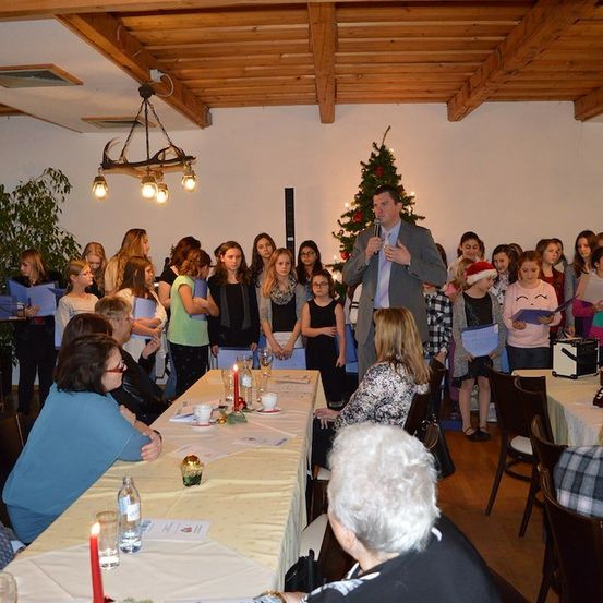 Eine Gruppe von Menschen ist in einem Raum für eine Weihnachtsveranstaltung versammelt. Ein Mann steht und spricht in ein Mikrofon. Ein mit Lichtern geschmückter Weihnachtsbaum steht hinter dem Mann. Die Menschen stehen und hören dem Mann zu. Es gibt Tische mit Papieren und Kerzen.
