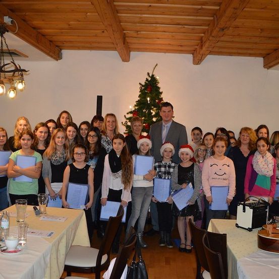 Eine Gruppe junger Mädchen und Frauen posiert für ein Foto in einem Raum mit einem Weihnachtsbaum und Dekorationen. Sie halten Urkunden in den Händen und einige tragen Mützen. Ein Mann in einem Anzug steht vor ihnen.