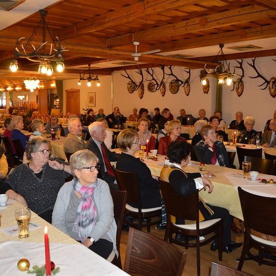 Eine Gruppe von Menschen sitzt an Tischen in einem Restaurant, einige mit Brillen und Schals, mit Kerzen und Gläsern auf den Tischen und Geweihen an der Wand.