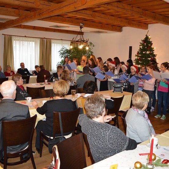 Eine Gruppe von Menschen versammelt sich in einem Raum für eine Weihnachtsveranstaltung, einige sitzen an Tischen und andere stehen vor einem Weihnachtsbaum und singen. Der Raum ist mit Weihnachtslichtern und Ornamenten geschmückt.