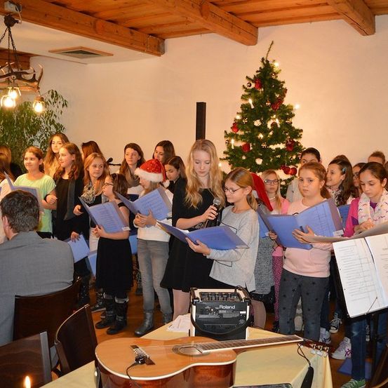 Ein Chor junger Mädchen in Winterkleidung singt Weihnachtslieder in einem für die Weihnachtszeit geschmückten Raum, mit einem Weihnachtsbaum und einem Mann, der Gitarre spielt, im Hintergrund.