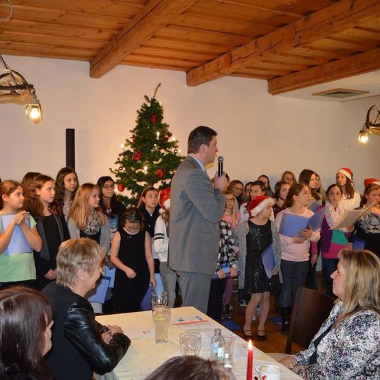 Eine Gruppe von Kindern und Erwachsenen versammelt sich in einem Raum mit einem Weihnachtsbaum, singt und hört einem Mann zu, der in ein Mikrofon spricht.