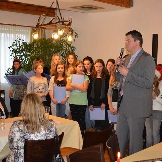 Ein Mann in einem Anzug steht vor einer Gruppe junger Mädchen mit Papieren und hält wahrscheinlich eine Rede. Dahinter sitzt eine Frau am Tisch. Im Hintergrund sind ein Weihnachtsbaum und ein Lautsprecher zu sehen.