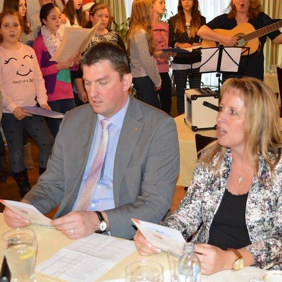 Ein Mann und eine Frau sitzen mit Papieren an einem Tisch, während ein Chor junger Mädchen im Hintergrund singt. Eine Frau spielt Gitarre und eine andere hält ein Notenpult.