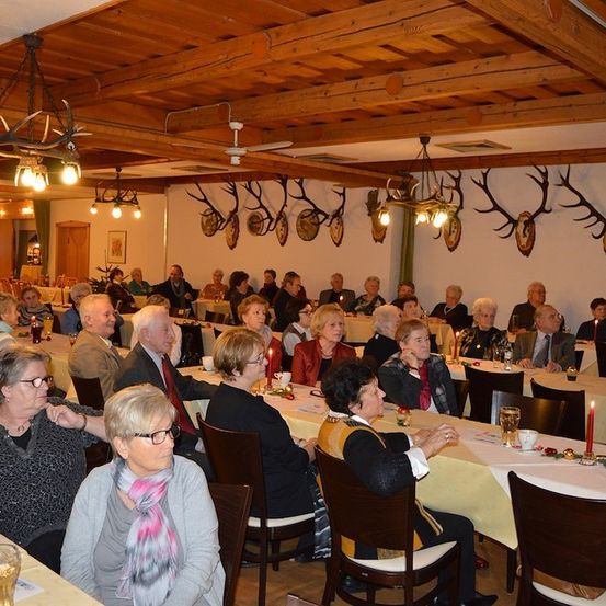 Eine Gruppe älterer Menschen versammelt sich in einem Speisesaal mit Holzbalken, Hirschgeweihen an der Wand und Kerzen auf den Tischen.