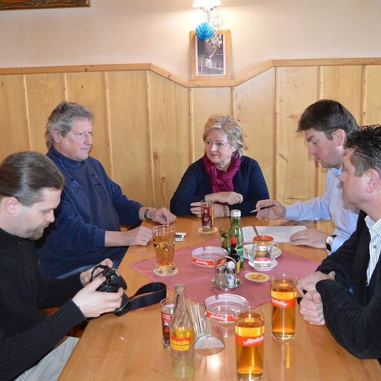 Fünf Personen sitzen um einen Holztisch herum, vermutlich in einem Restaurant. Sie führen ein Gespräch, wobei einige Biergläser halten. Der Tisch ist mit verschiedenen Gegenständen geschmückt, darunter Flaschen, ein Salzstreuer und eine rosa Matte. Die Wand hinter ihnen ist aus Holz, und ein Bilderrahmen ist daran befestigt.