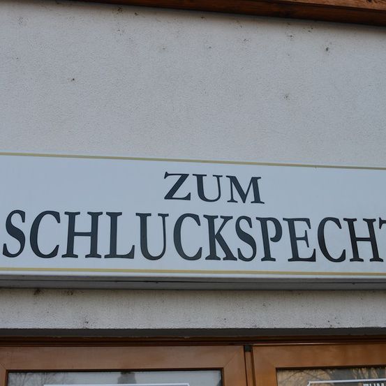 Ein weißes Schild mit schwarzem Text, das an einer Wand über einer Holztür befestigt ist. Das Schild trägt den Text 'ZUM SCHLUCKSPECHT'.