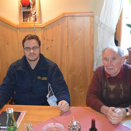 Zwei Männer sitzen an einem Tisch in einem Restaurant, einer trägt eine Polizeijacke und der andere ein kastanienbraunes Sweatshirt. Dahinter eine Holzwand, eine Topfpflanze und ein gerahmtes Bild.
