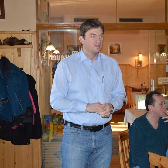 Ein Mann in einem hellblauen Hemd und Jeans steht in einem Raum und spricht zu einem anderen Mann, der an einem Tisch sitzt. Der Raum hat Holzwände und eine Glastür.