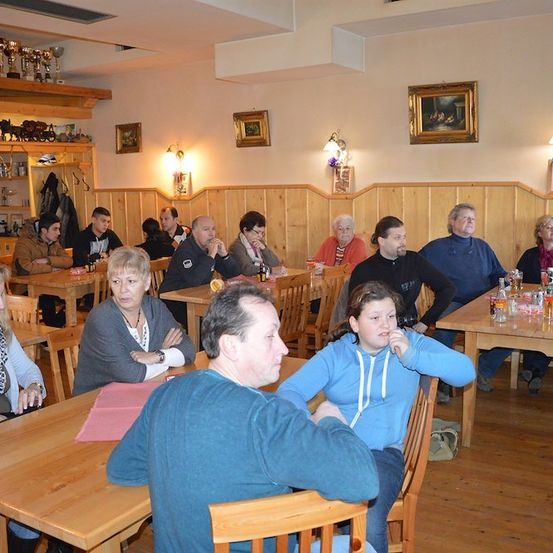 Eine Gruppe von Menschen sitzt an Tischen in einem Restaurant mit Holzwänden und Regalen mit Trophäen und Bilderrahmen.