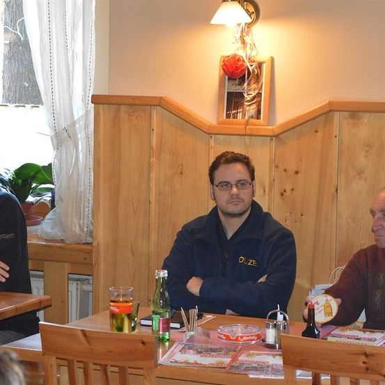 Drei Männer sitzen an einem Tisch in einem Restaurant. Der Mann in der Mitte trägt eine blaue Jacke mit dem Wort 'DUZE' darauf. Der Mann rechts hat eine Flasche und einen Teller vor sich. Hinter ihnen ist eine Wand mit einem Bilderrahmen und einer Lampe.