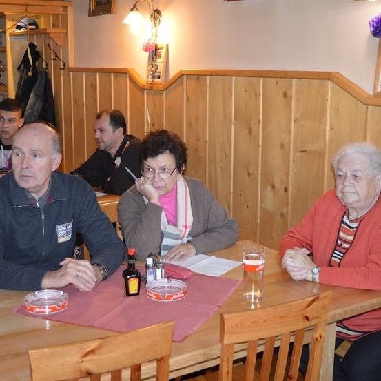 Eine Gruppe von Menschen sitzt in einem Restaurant an einem Tisch, wobei eine Frau einen Stift und Papier hält und eine andere Frau sie ansieht. Auf dem Tisch stehen eine Flasche, ein Glas und ein Aschenbecher. Hinter ihnen sitzt ein Mann mit einer Jacke, die an einem Kleiderständer hängt.