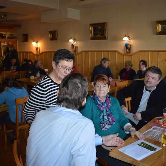 Eine Gruppe von Menschen sitzt in einem Restaurant an Tischen, wobei eine Frau in einem grünen Pullover und einer Schürze einen Mann in einem blauen Hemd ansieht.