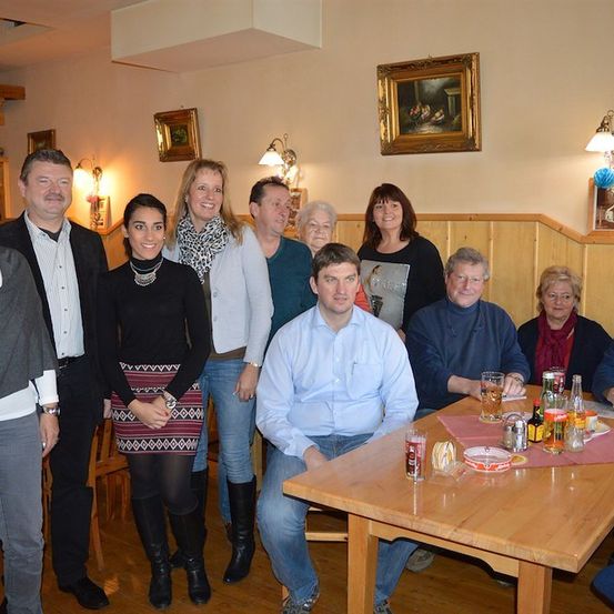 Eine Gruppe von Menschen posiert für ein Foto in einem Restaurant. Sie stehen und sitzen um einen Tisch mit Getränken und Essen. Der Raum hat Holzwände und eine Decke mit Lampen.
