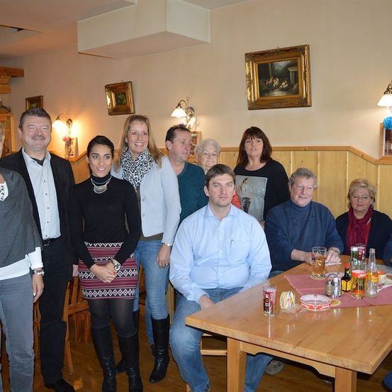 Eine Gruppe von Menschen posiert für ein Foto in einem Raum mit Holzwänden, einem Tisch und verschiedenen Gegenständen darauf. Einige stehen und andere sitzen. Es gibt Bilderrahmen an der Wand.