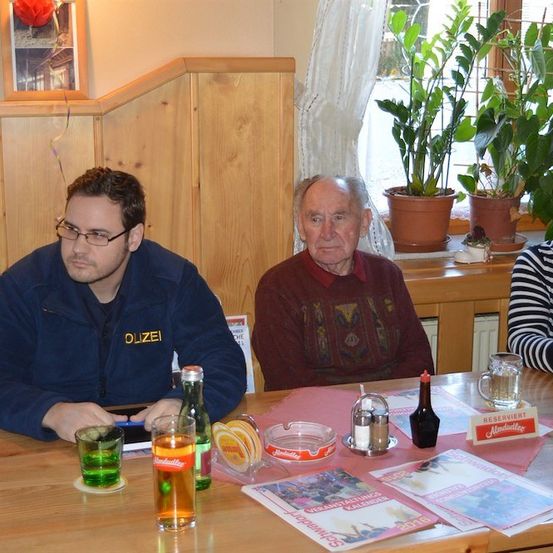 Drei Personen sitzen an einem Tisch mit Getränken und Essen. Ein Mann mit Brille schaut auf sein Handy, ein älterer Mann in einem roten Pullover schaut in die Kamera und eine Frau in einem gestreiften Shirt schaut weg.