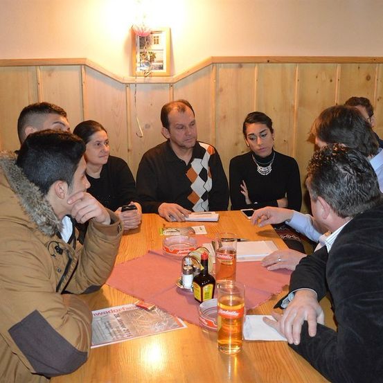 Eine Gruppe von Menschen sitzt um einen Holztisch herum und führt ein Gespräch. Der Tisch ist mit Biergläsern, Flaschen und verschiedenen Gegenständen bestückt. Der Raum hat Holzwände und ein Bildrahmen an der Wand.