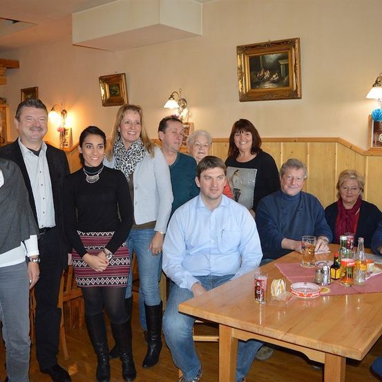 Eine Gruppe von Menschen, darunter Männer und Frauen, versammelt sich in einem Raum, möglicherweise für ein Familientreffen. Sie stehen und sitzen um einen Tisch mit Getränken und Essen. Der Raum hat Holzwände und eine Decke mit Lampen.