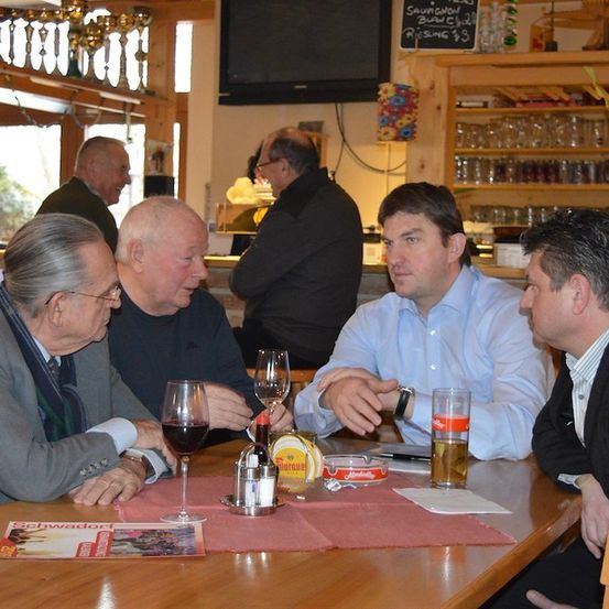 Vier Männer sitzen in einem Restaurant um einen Tisch, zwei von ihnen halten Weinglas in der Hand. Ein Mann trägt eine Uhr, und ein anderer hat ein Bier vor sich. Hinter ihnen sind eine Tafel und ein Fernseher zu sehen.