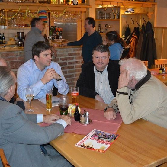 Fünf Männer sitzen in einer Bar um einen Tisch, zwei von ihnen trinken Bier, während die anderen in ein Gespräch vertieft sind. Ein Kalender liegt auf dem Tisch.