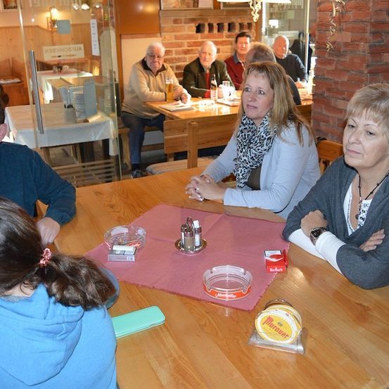 Eine Gruppe von Menschen sitzt in einem Restaurant um einen Tisch herum, wobei zwei Frauen in die Kamera schauen. Der Tisch hat ein rosa Tuch, Zigaretten und einen Senfbehälter.