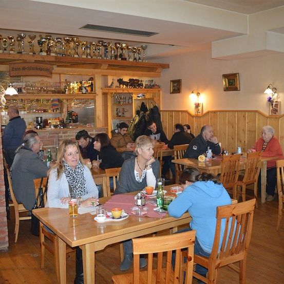 Eine Gruppe von Menschen sitzt an Tischen in einem Restaurant mit Holzwänden und -böden. Hinter der Bar befinden sich Trophäen auf einem Regal.