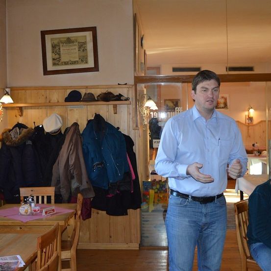 Ein Mann steht in einem Raum mit Holzwänden und spricht, während ein anderer Mann am Tisch im Hintergrund sitzt. Mäntel hängen an der Wand und ein gerahmtes Bild ist über ihnen angebracht.