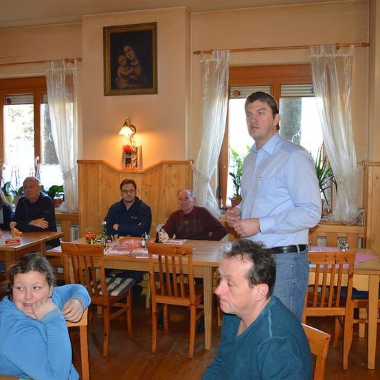 Eine Gruppe von Menschen sitzt an Tischen in einem Restaurant. Ein Mann steht und spricht zu ihnen.