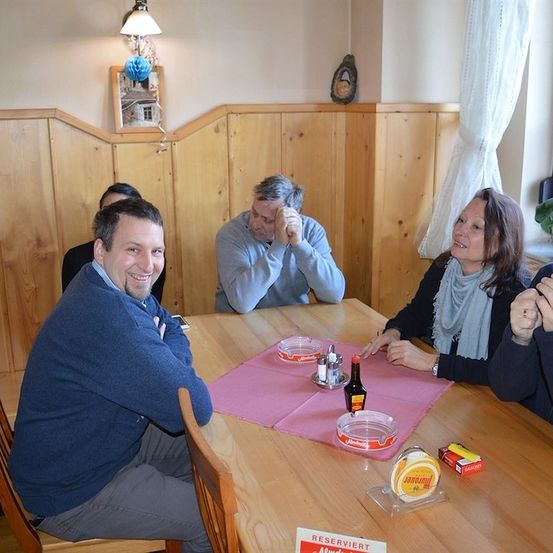 Eine Gruppe von Menschen sitzt um einen Tisch herum, lächelt und lacht. Ein Mann auf der rechten Seite raucht eine Zigarette. Der Tisch hat ein rosa Tuch, eine Flasche, eine Schachtel und einen Teller. Hinter ihnen ist eine Holzwand mit einem Bilderrahmen und einer Lampe.