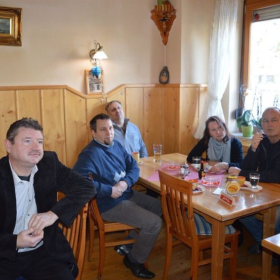 Fünf Personen sitzen an einem Tisch in einem hölzernen Raum, wobei eine Frau mit einer Schürze die Kamera ansieht. Auf dem Tisch stehen Gläser, Teller und Flaschen.