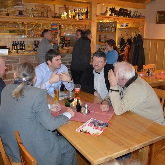 Eine Gruppe von Menschen sitzt in einer Bar um einen Tisch herum, einige unterhalten sich, während andere etwas betrachten. Auf dem Tisch stehen Flaschen und Gläser.