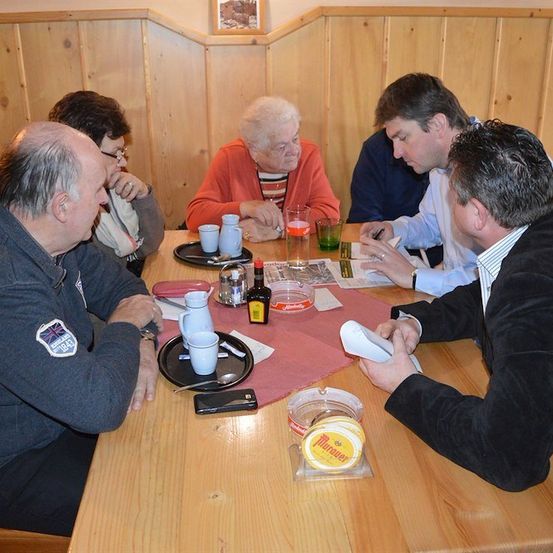 Eine Gruppe von Menschen sitzt um einen Holztisch in einem Restaurant, mit verschiedenen Gegenständen auf dem Tisch wie Tassen, Flaschen und eine Zeitung. Eine ältere Frau sitzt in der Mitte und ein Mann schreibt auf ein Stück Papier.
