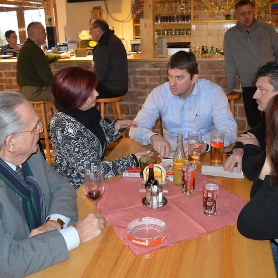 Eine Gruppe von Menschen sitzt in einer Bar um einen Tisch herum und führt ein Gespräch. Auf dem Tisch befinden sich Getränke und Snacks. Hinter ihnen ist eine Ziegelwand und eine Bartheke zu sehen.
