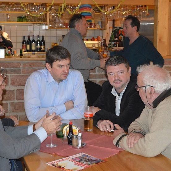 Eine Gruppe von Männern sitzt in einer Bar um einen Tisch herum, wobei einer der Männer hinter der Theke steht. Sie alle sehen sich an und scheinen ein Gespräch zu führen. Auf dem Tisch stehen Flaschen und Gläser, und eine Speisekarte ist darauf gelegt.