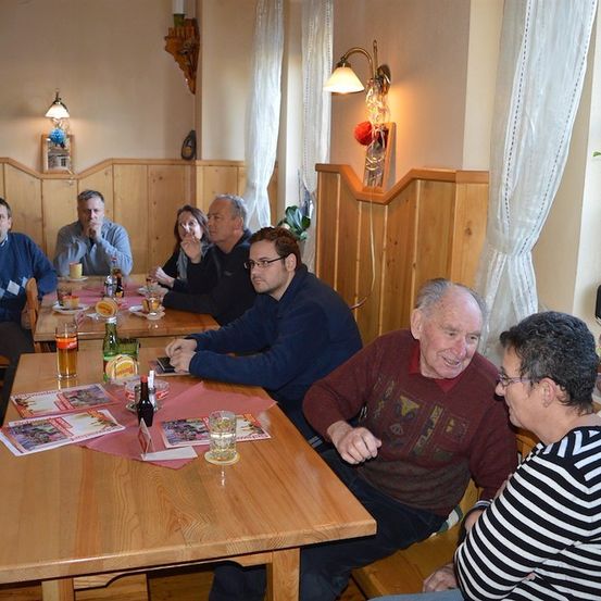 Eine Gruppe von Menschen sitzt an einem Tisch in einem Restaurant. Sie führen ein Gespräch. Der Tisch ist mit Getränken, Essen und Servietten gedeckt. Der Raum hat Holzwände und weiße Vorhänge.